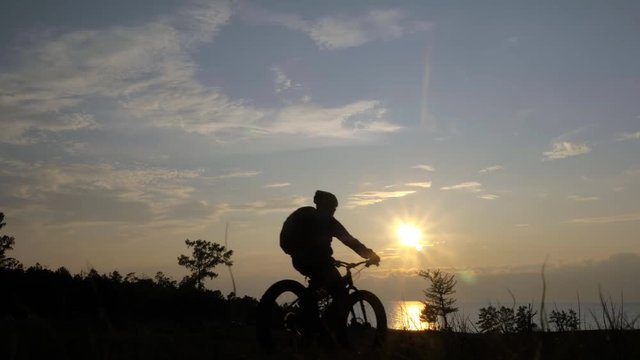 Fat bike also called fatbike or fat-tire bike in summer riding in the grass. The athlete passes in a frame silhouette against a beautiful sunset on the sea. Everything happens in deep grass.