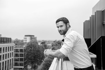 Handsome hunky man in tuxedo, unbuttoned shirt and tie stands on hotel balcony, drinking a beverage