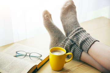 Woman resting keeping legs in warm socks on table with morning coffee and reading book - 216692232