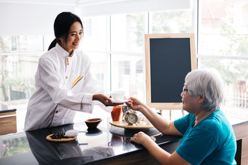Young Asian female barista making and giving coffee to senior old customer. Retired woman receiving coffee at cafe restaurant
