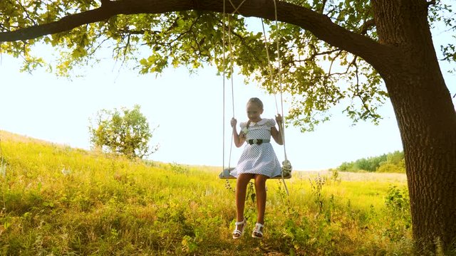 Beautiful girl with long hair swings on swing under summer oak in white dress.