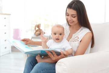 Young mother with her cute baby girl reading book in armchair at home