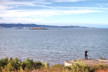 A picture of a lone man taking a photograph of the now deserted island of Inchmickery. The photograph was taken on Cramond Island overlooking the Firth of Forth near Edinburgh, Scotland, UK.