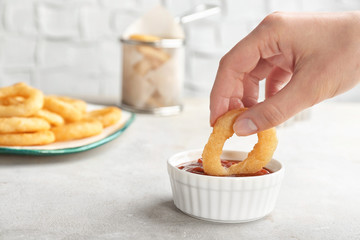 Woman dipping tasty onion ring into bowl with ketchup on table
