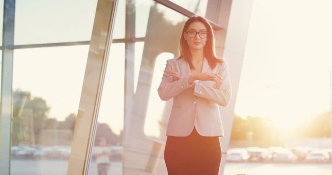 Portrait shot of the charming Caucasian businesswoman standing outdoor glass modern office building, turning her head to the camera with a smile and crossing hands in the sunlight.