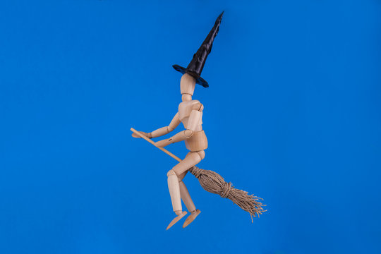 Jointed wooden doll dressed as a witch riding a witches broom wearing a black witches hat on blue background 