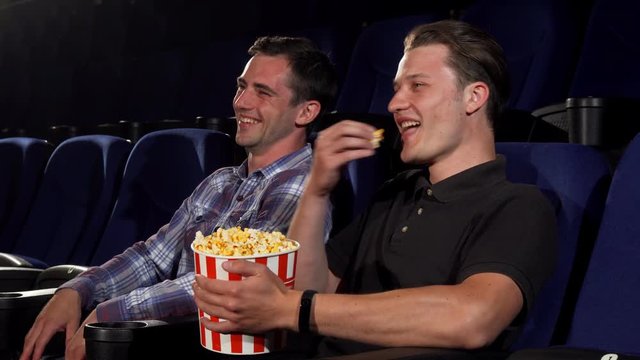Young handsome man laughing joyfully talking to his friend while watching comedy movie at the cinema. Cheerful male friends eating popcorn at the movie theatre. Entertainment concept.