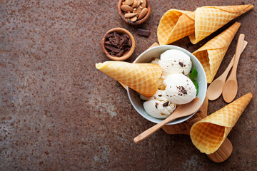 Vanilla ice cream scoops with pieces of chocolate bar and waffle cones on brown background.