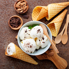 Vanilla ice cream scoops with pieces of chocolate bar and waffle cones on brown background.