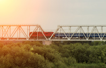 Passenger train rides along the railway bridge over the river, the sun