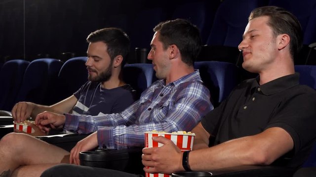 Cheerful young men talking, eating popcorn, enjoying watching movies at the cinema together. Male friends relaxing at the movie theatre. Friendship, lifestyle, happiness concept.