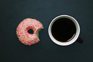 Bitten donut in pink glaze and cup of coffee on dark background, view from above