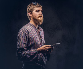 Redhead bearded hipster guy dressed in an old-fashioned shirt holds a tablet computer on a dark background.