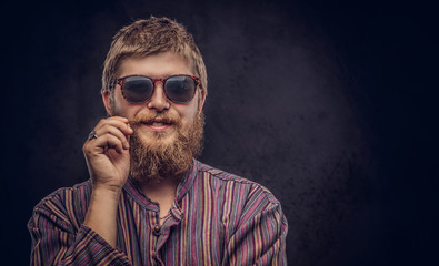 Happy hipster guy wearing sunglasses dressed in an old-fashioned shirt correct his mustache on a dark background.