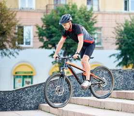 Fototapeta premium Handsome man bicyclist in cycling clothing and protective gear riding down concrete stairs in city urban area. Sportsman performing tricks on bike, training for contest. Concept of healthy life