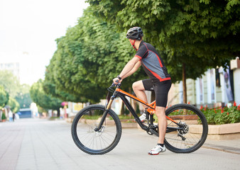 Side view of guy cyclist in cycling clothes and helmet looking away getting ready to cross city street on bicycle. Sportsman shooting advertising sport campaign. Man exercising, training outdoors