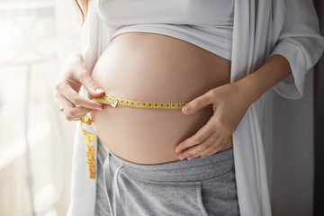 Close-up shot of pregnant happy woman holding ruler and measuring size of belly, making notes during pregnancy period, looking after health and baby condition, standing near window at home