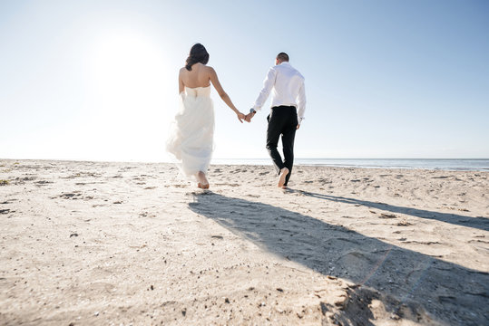 Wide angle photo of beautiful wedding couple bride and groom at wedding day is walking along the beach at sunny evening