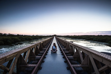 Young fashionable dressed woman is sitting on a steel brige urban design by the lake with sunset