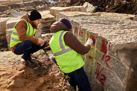 Back view of two industrial workers wearing reflective jackets marking  granite on mining worksite outdoors