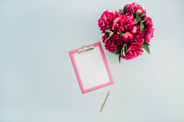 Minimalist flat lay office table desk with clipboard, pink peony flower bouquet on blue background. Top view work concept mock up.