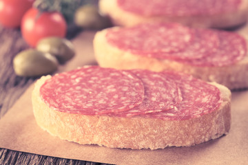 Salami slices on ciabatta bread with green olives and cherry tomato in the back (Selective Focus, Focus on the front of salami slices on first sandwich) (Digitally Altered: Toned Image)