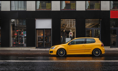 Yellow car is parked at asphalt road in the city center in the rain. Storefront - obrazy, fototapety, plakaty