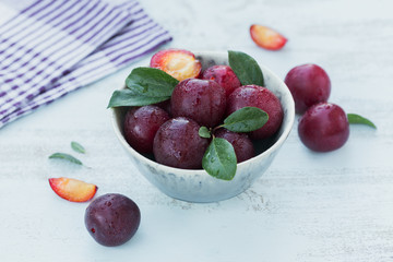 Fresh plums with leaves in a bowl on white rustic wooden table background.