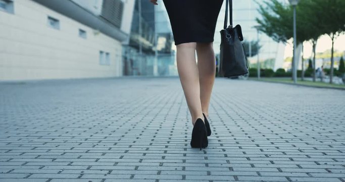 Rear of the female legs in the short skirt and shoes with heels walking on the street.