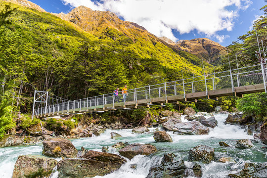 New Zealand tramping people crossing river bridge. Hikers couple backpackers walking hiking together with backpacks on Routeburn Track trail path.
