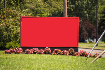 Large blank red sign board on green grass with small flowers at its base and blurred out tree...