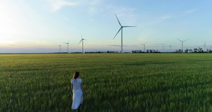 Beautiful girl walking on a green wheat field with windmills for electric power production