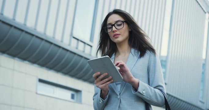 Portrait of the good looking young businesswoman thinking while typing and taping on the tablet device outside the office building center.