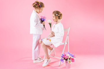 Twins girls in light clothes with bouquets of flowers posing near a chair on a pink background.