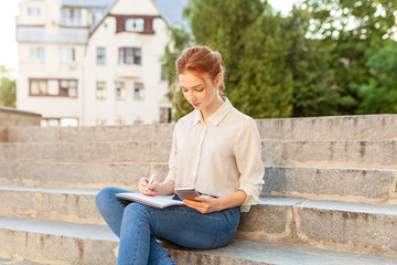 Obraz premium beautiful young redhead girl with freckles sitting on a stairs and making notes in a notebook. Portrait of a student. Remote education. Homework