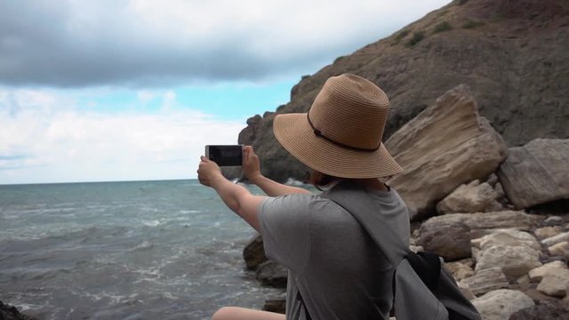 Woman with a hat takes pictures of the sea on her mobile phone on the beach
