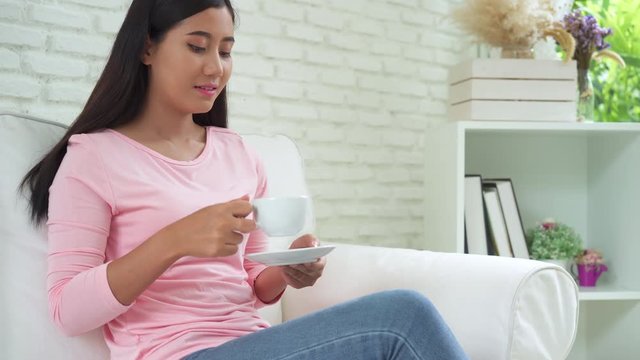 Cheerful asian young woman drinking warm coffee or tea enjoying it while sitting in her living room at home. Attractive happy asian woman holding a cup of coffee.