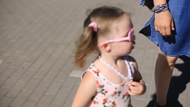 a little two-year-old girl with blonde hair and sunglasses is walking on the sidewalk tiles in a Park Sunny bright day in a dress.