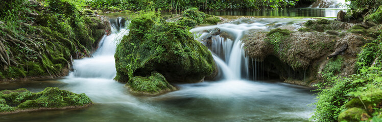 Panoramic view of small waterfalls streaming into small pond in green forest in long exposure - 216750678