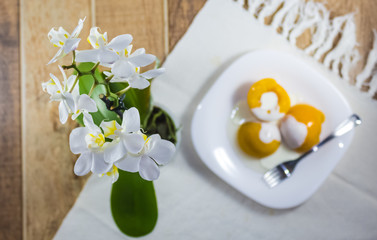 Peach in syrup with cream milk, dessert served on a white dish on a table decorated with vase of orchids.