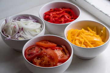 top diagonal view of four cups of sliced vegetables in white bowls on white counter top: onions, red peppers, tomatoes and yellow peppers