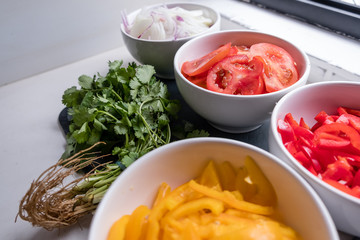 top diagonal view of coriander and four cups of sliced vegetables in white bowls on white counter top: onions, bell peppers, tomatoes. Ingredient for traditional brazilian dish called moqueca