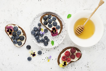 Light summer snack toasts with ricotta, honey, blueberries, raspberries and edible flowers of garden violets on dark-colored whole wheat bread on a light background. Top View.