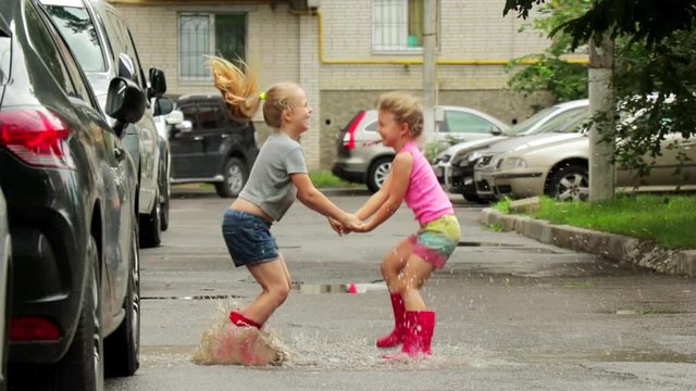 SLOW MOTION: Two little girls in pink rubber boots have fun jumping in a puddle. nearby are the cars in the yard of a large new brick house. Children are happy.