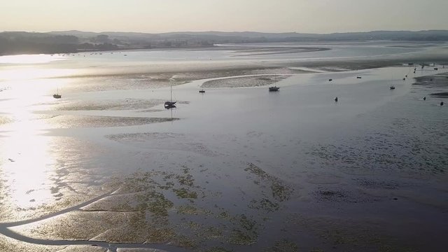 Flying over the beach of Lympstone with its multiple boats docked off the shore. The sun is setting and the glow is reflecting in the water giving it a golden brown tone. Tourists visit daily.