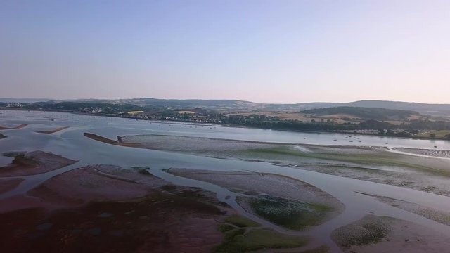Flying over the beautiful landscape of Lympstone beach. The sky is clear, the hills and mountains in the background, and the water has patches of sand formations.