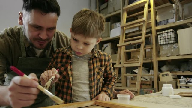 Medium shot with low angle of happy bearded father in apron showing little boy how to put varnish on wooden picture frame