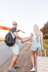 A loving couple walks along the road with a guitar and suitcases and catches a car hitchhiking.