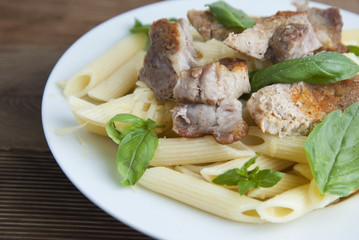 Cooked penne pasta isolated in white round plate, with basil leaves and pork meat, over wooden table, decorated with basil leaves. Top view.