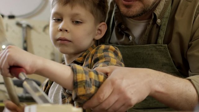 Medium shot of cheerful male carpenter helping cute little boy using mallet and chisel to carve wood in workshop